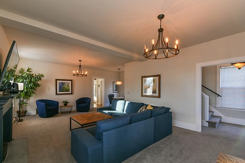A spacious living area features a comfortable navy blue sectional sofa, complemented by two cushioned chairs. A wooden coffee table sits at the center, while artwork adorns the walls. Soft lighting from multiple chandeliers creates a welcoming environment.