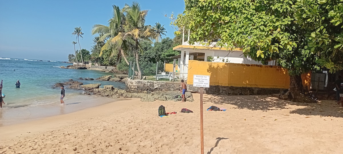 A sandy beach area is shown, framed by palm trees. The clear water laps at the shore, while a yellow building is partially visible in the background. A sign is positioned in the foreground, and beachgoers can be seen enjoying the water.