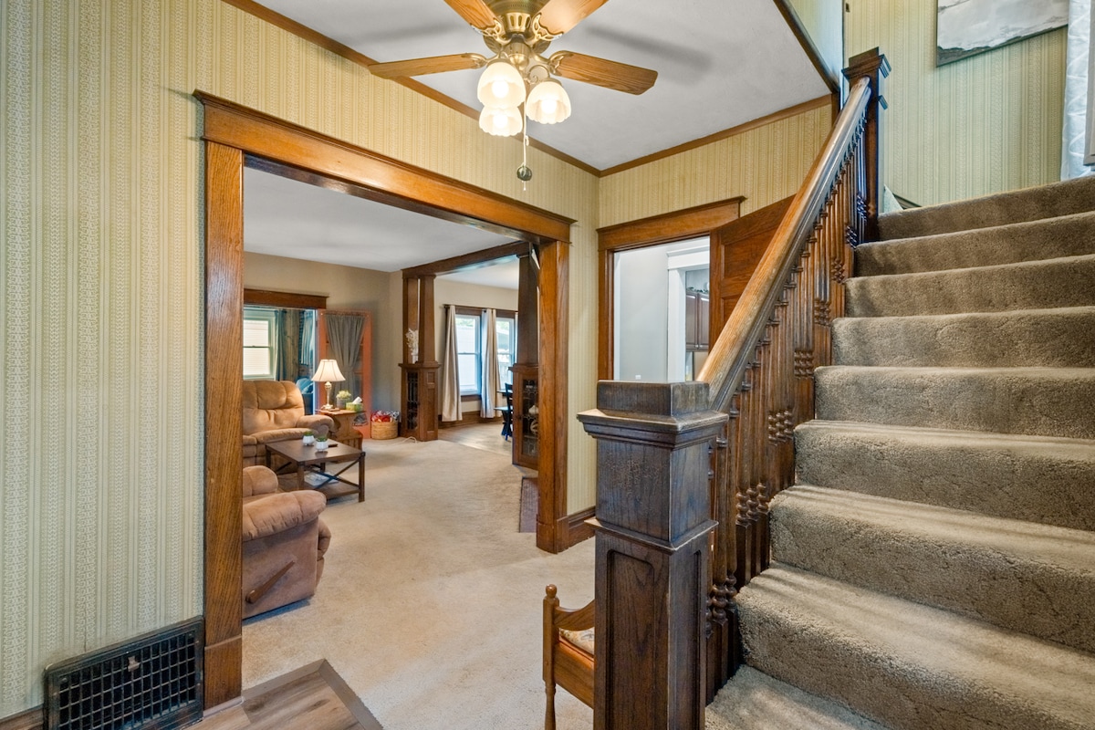A warm entryway features a staircase leading to the upper level, accompanied by wooden railings and a light fixture. The living area is visible through an open archway, revealing comfortable seating and natural light from large windows.