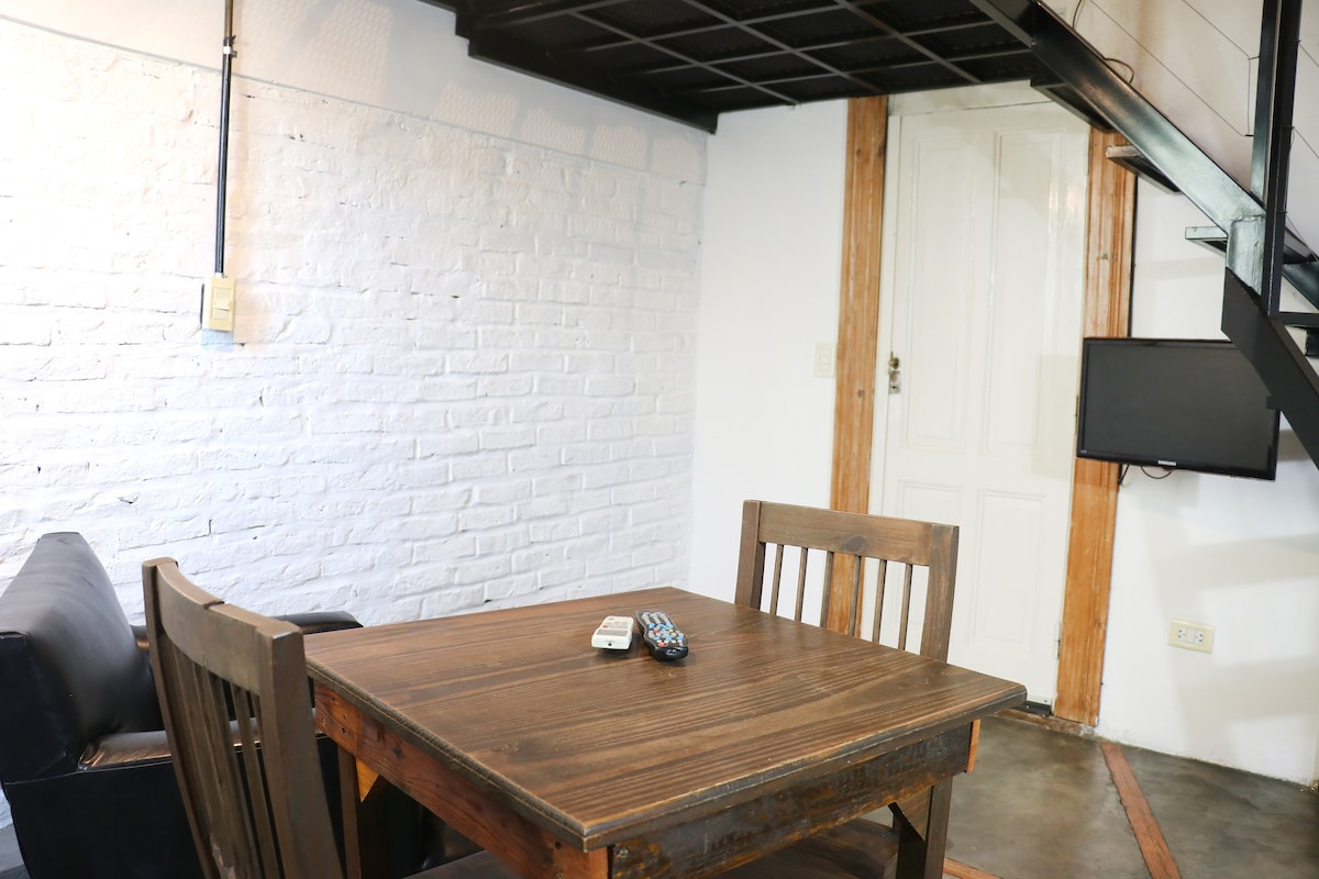 A cozy sitting area is highlighted by a wooden table with two matching chairs. A remote control is placed on the table. The wall features exposed white brick, while a staircase leads to an upper level. A small television is mounted on the adjacent wall.