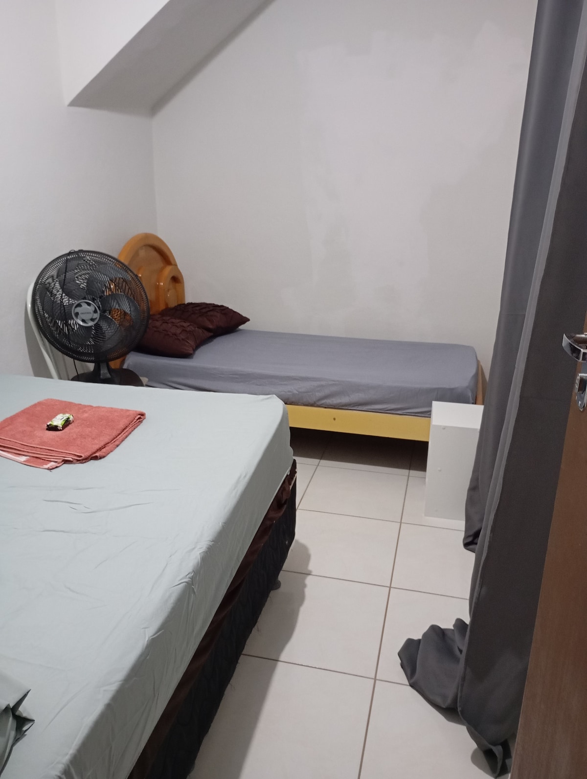 A small bedroom is presented with two beds, one with a light blue cover and the other with a gray cover. A fan is positioned nearby, and a towel set is neatly arranged on the bed. The walls are painted white, enhancing the room's simplicity.