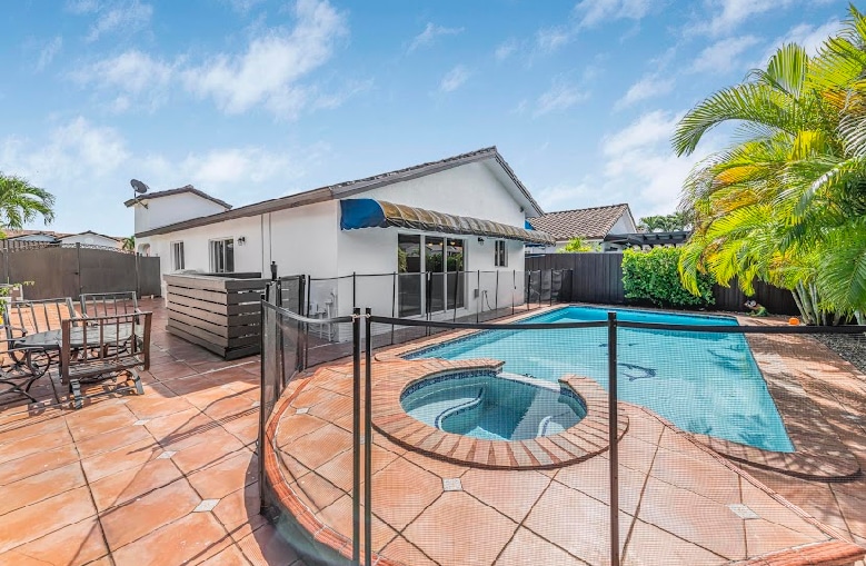 A spacious outdoor area features a swimming pool and a hot tub, surrounded by a protective fence. The patio is tiled and includes a dining set, complemented by lush greenery and palm trees that add to the serenity of the space.