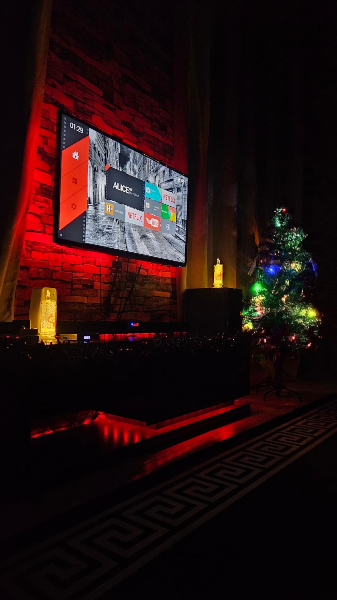 A modern entertainment setup is highlighted by a large screen television mounted against a stone wall. Colorful lights adorn a nearby decorated Christmas tree, creating a festive ambiance. A low console table features dim lighting, enhancing the cozy atmosphere.