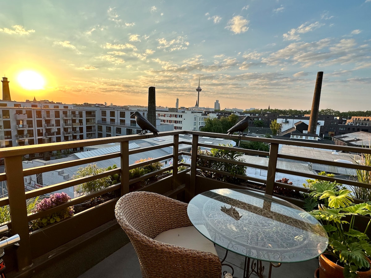 A balcony view showcases a vibrant sunset over the cityscape of Ehrenfeld. A glass table with a wicker chair is positioned in the foreground, surrounded by potted plants. Distant landmarks like a television tower are visible, with soft clouds adding texture to the sky.