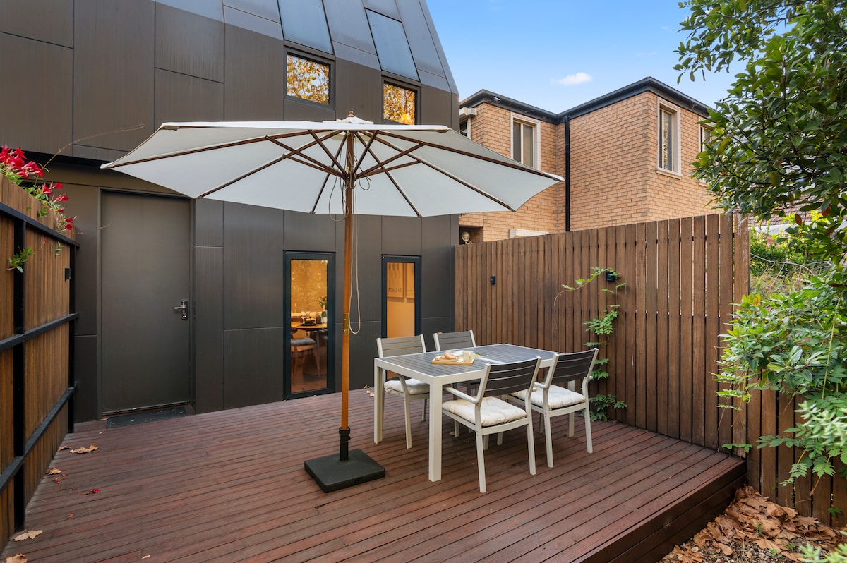 A private patio area is presented with a wooden deck and a dining table surrounded by four chairs. An umbrella provides shade for the table. Lush green plants and flowers accentuate the space, while a backdrop of the house's modern architecture is visible.