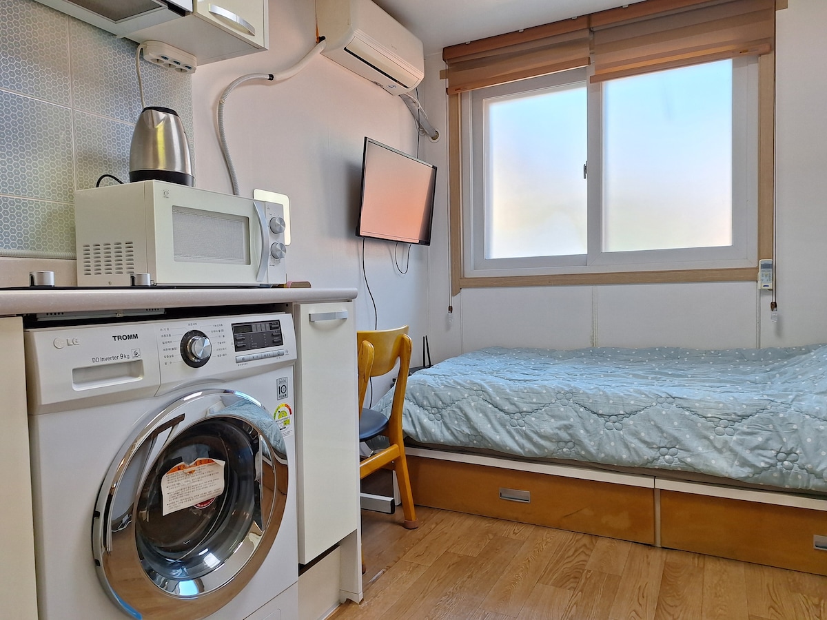 A compact living space features a spacious bed with a light quilt, complemented by a simple chair and a mounted television. A washing machine is integrated into the kitchen area, alongside a microwave and a kettle. Natural light enters through a window with wooden blinds.
