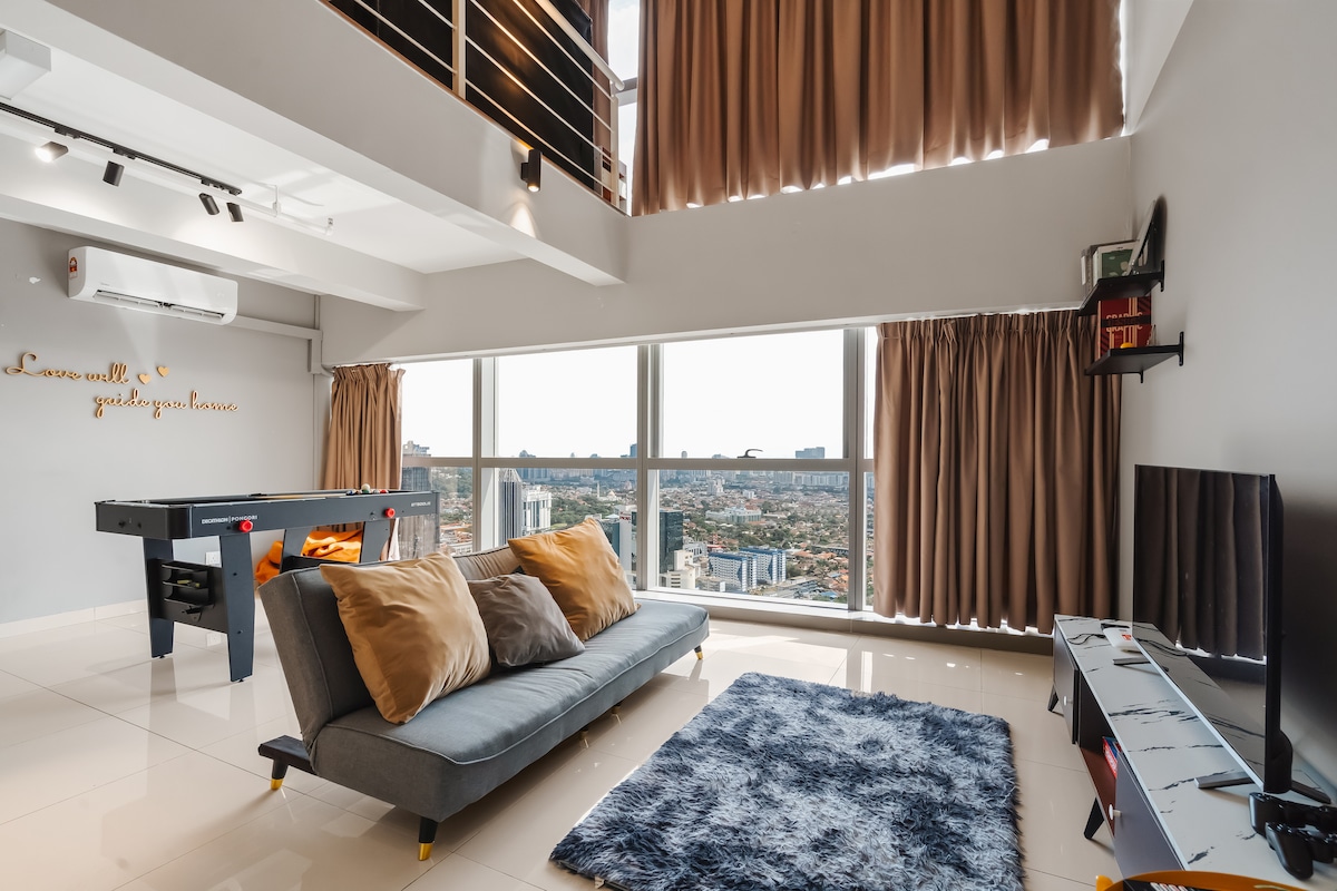 A spacious living area features large windows offering a city view, allowing natural light to fill the room. A comfortable gray sofa is adorned with decorative cushions, while a soft rug lies beneath. An entertainment setup with a TV and game table adds functionality.