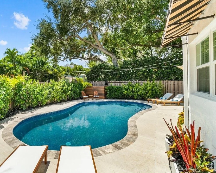 A private heated pool is surrounded by lush greenery and tropical plants. Comfortable lounge chairs are positioned nearby, providing a relaxing sunbathing area. Soft lighting strings are draped overhead, enhancing the outdoor ambiance. Sunlight reflects off the water, inviting guests to enjoy the serene environment.