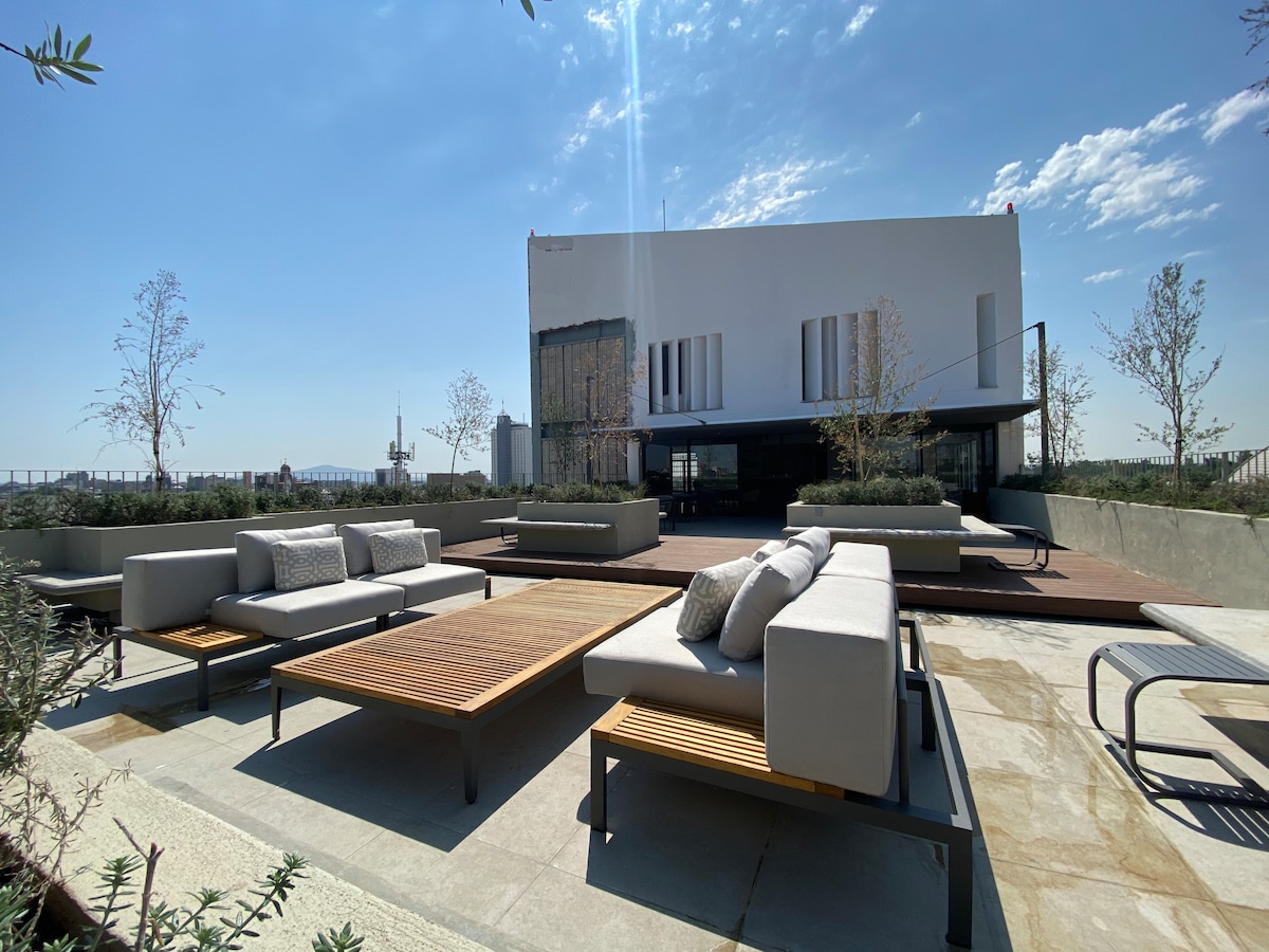 An outdoor terrace features modern seating arrangements with plush cushions, providing ample space for relaxation. Wooden tables are positioned among low greenery, offering a refreshing atmosphere. The background showcases a contemporary building with large windows, allowing natural light to fill the area.