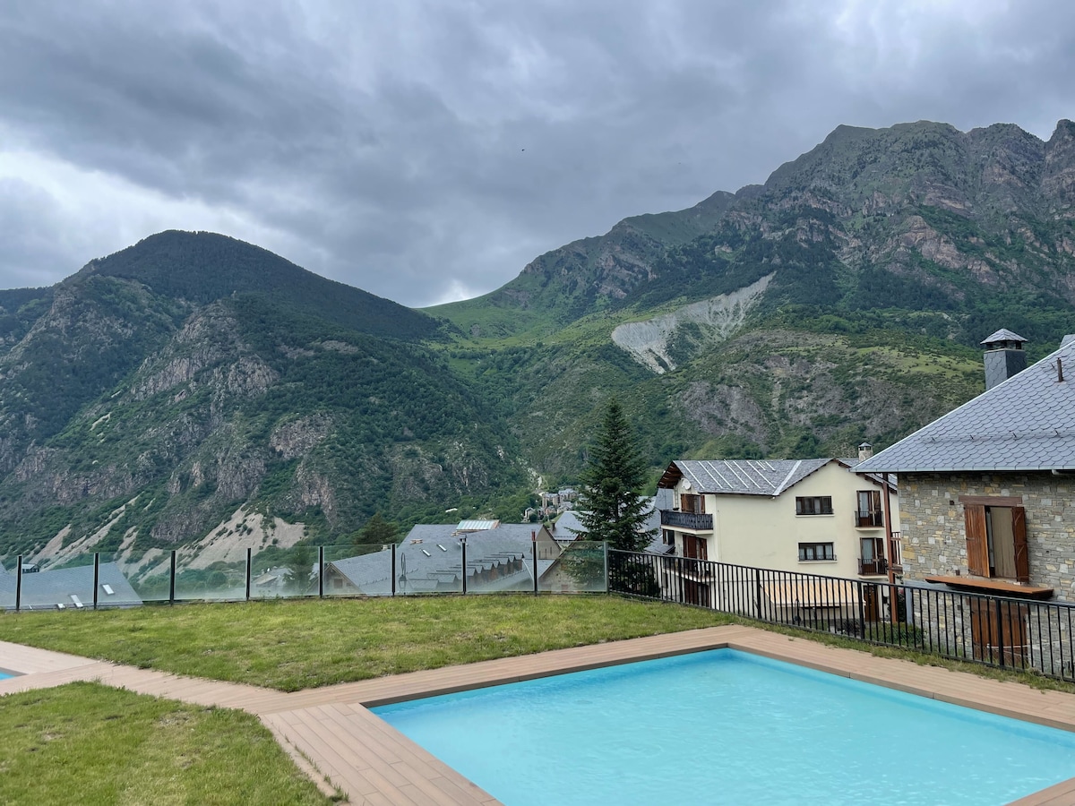 A tranquil outdoor area features a clear swimming pool framed by a wooden deck. Lush green mountains rise in the background, partially shrouded by clouds, and several residences are visible with a combination of stone and wooden facades.