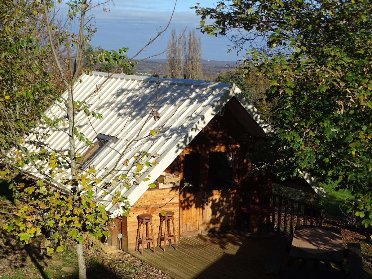 A wooden cabin is nestled among trees, featuring a sloped metal roof. Bar stools are positioned on a deck area, inviting relaxation. The landscape in the background displays greenery and distant trees under a calm sky.