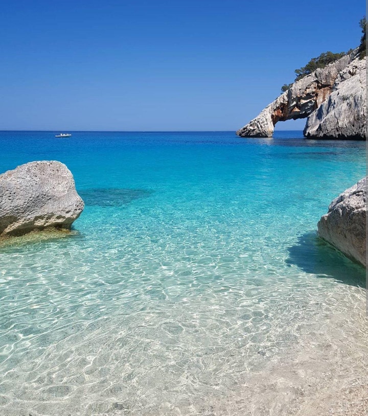 A clear blue sea stretches toward the horizon, featuring calm waters that reveal sandy shores beneath. A rocky formation arches gracefully over the water, accompanied by smooth stones along the shoreline. The bright sky enhances the tranquil scene, inviting relaxation and exploration.