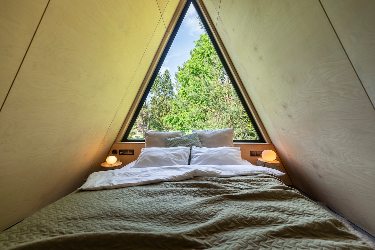 A spacious bed is positioned in a triangular room, framed by large windows that offer views of surrounding greenery. Soft bedding is arranged neatly, with two lamps placed on either side of the bed, providing gentle illumination.