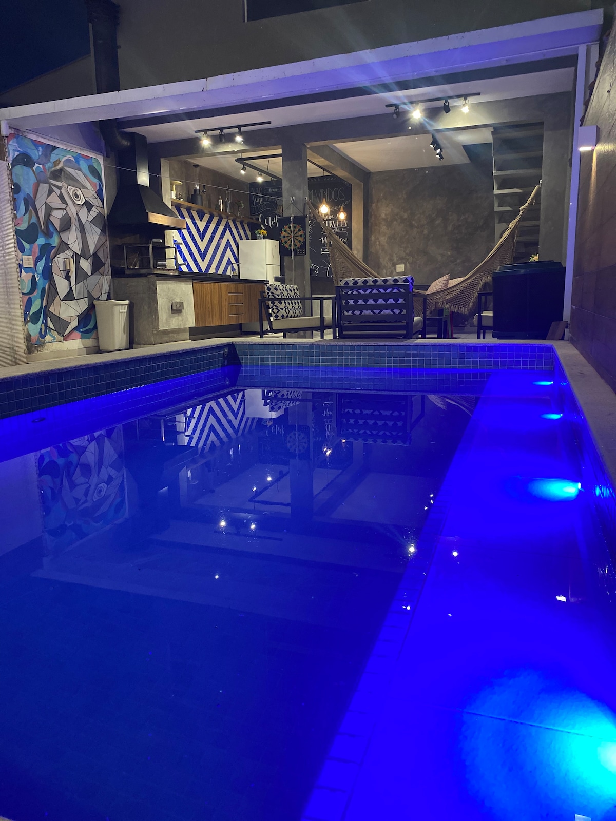 A tranquil pool area is highlighted by softly illuminated water, reflecting the modern design of the surrounding space. Colorful artwork adorns the walls, and seating arrangements are positioned nearby. The open kitchen is visible in the background, featuring sleek cabinetry and fixtures.