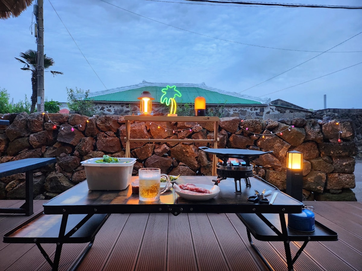 An outdoor dining space is set up with a grill and food items on a table. Decorative lights hang along a stone wall. A warm glow from lamps adds to the ambiance, while a palm tree neon sign illuminates the scene against a twilight sky.