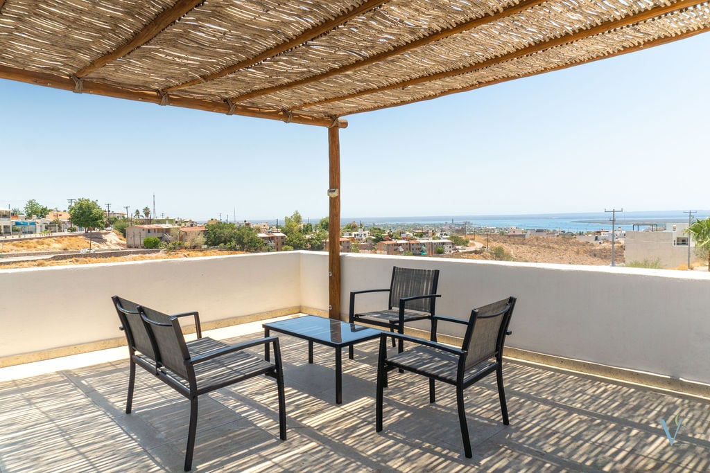 A private terrace is displayed, featuring a shaded pergola and a seating area with four black chairs around a glass table. The view includes distant hills and the shimmering bay, creating a calming outdoor space.