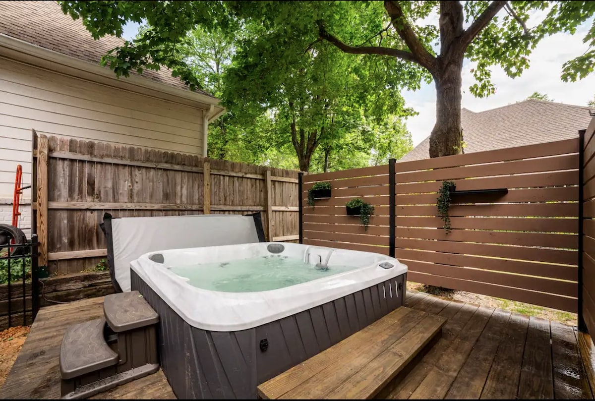 A private hot tub is nestled within a fenced wooden patio area, surrounded by lush greenery and mature trees. The tub features steps for easy access and is covered by a protective hard top when not in use.