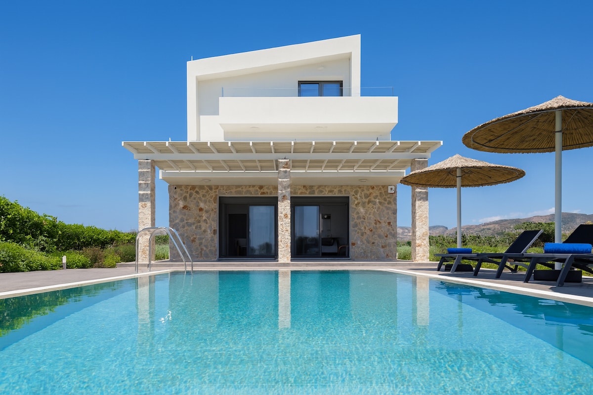 A modern villa showcases clean lines and a combination of stone and white façade against a vibrant blue sky. A private pool reflects sunlight, while lounge chairs and umbrellas are arranged around the pool area, inviting relaxation in a scenic outdoor environment.