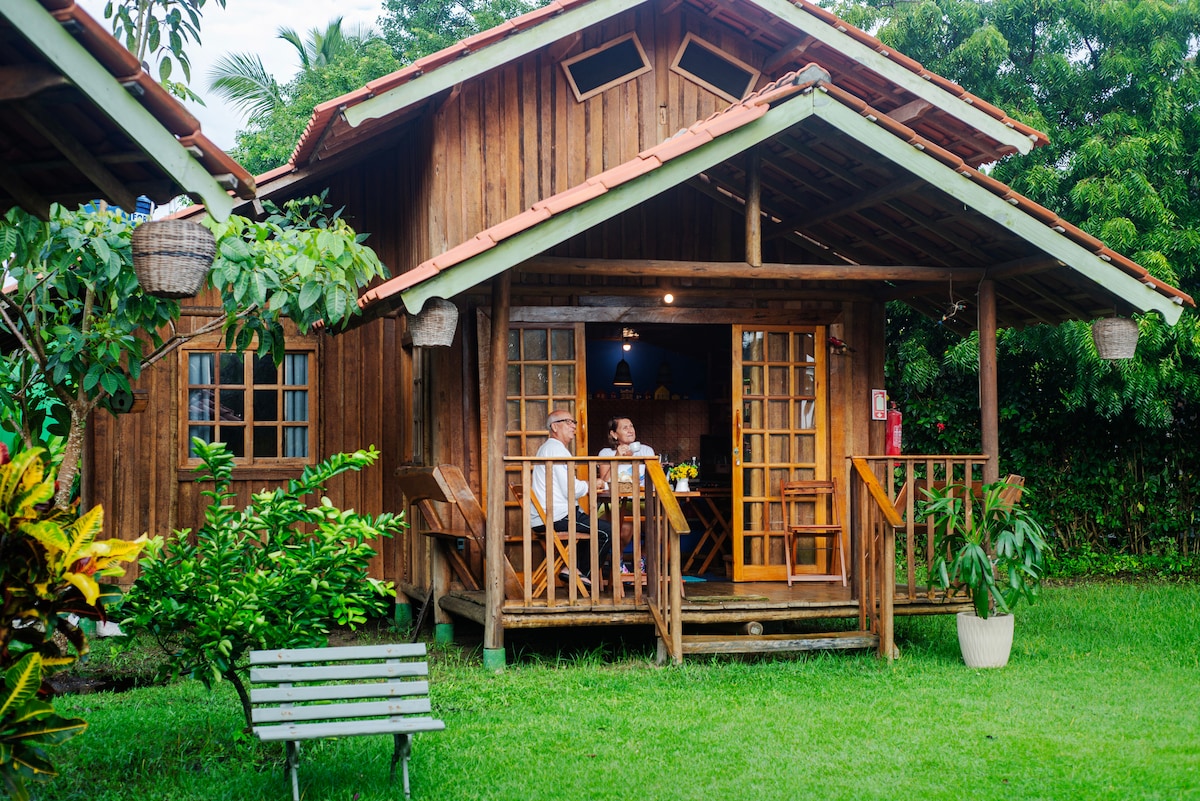 A quaint wooden cabin is nestled among greenery, featuring a welcoming porch with a railing and wooden chairs. Soft light emanates from the interior, while large windows provide ample natural light. A small bench sits in the grass nearby, enhancing the outdoor space.