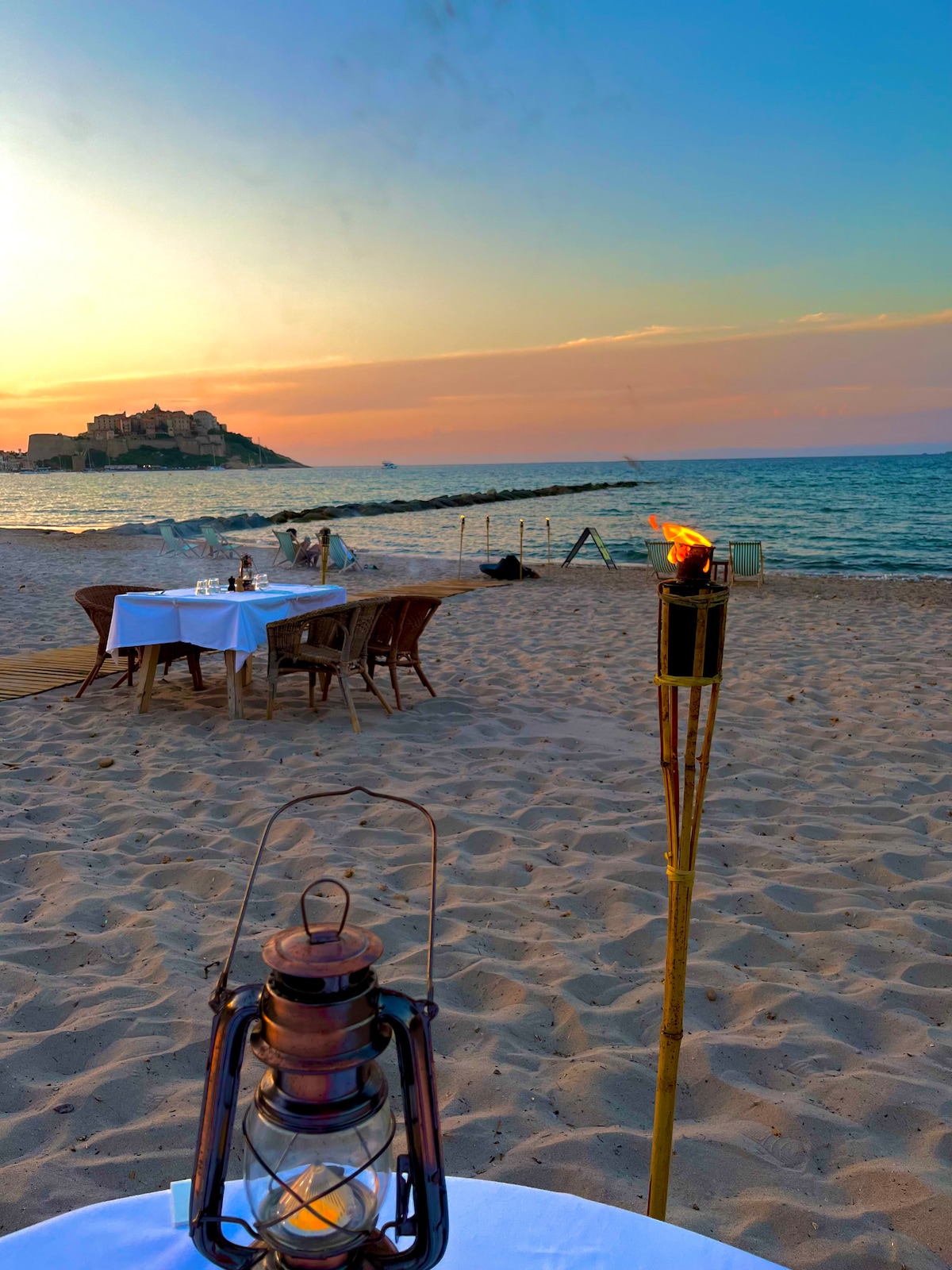 A beachside table is set for dining, surrounded by wooden chairs on soft sand. A tiki torch with a flickering flame stands nearby, providing a gentle glow. In the background, the calm sea meets the shore, and a distant hill is visible under a colorful sunset.