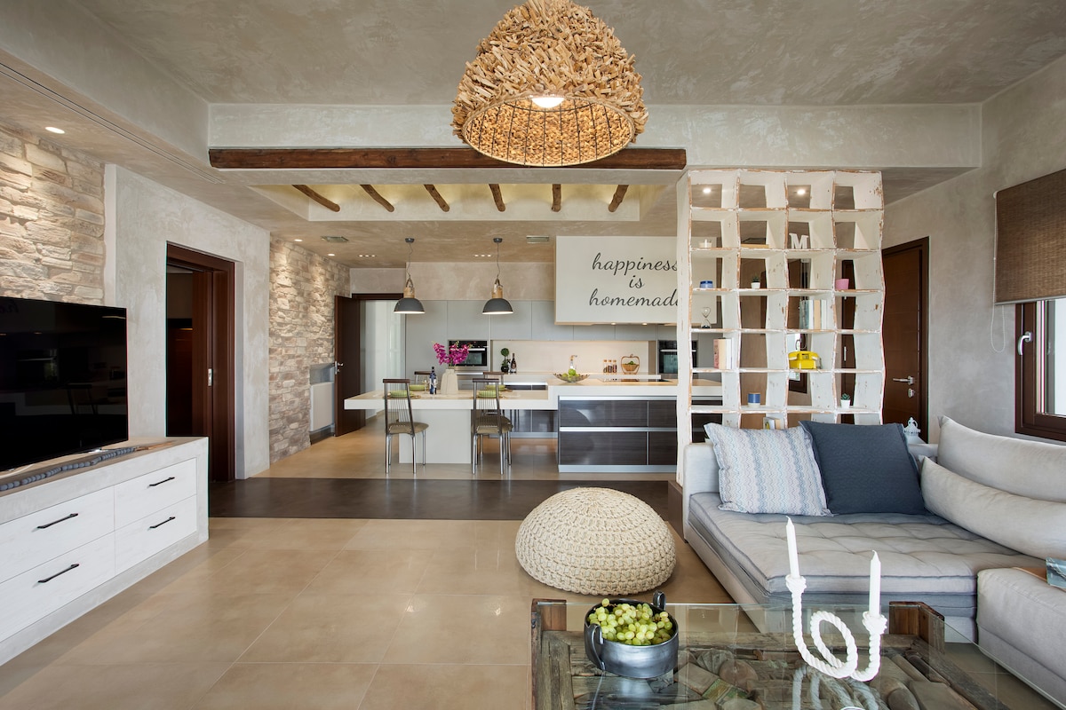 The interior showcases a modern open-plan living area with natural stone accents. A spacious sofa is centered in the room, complemented by a glass coffee table. A unique woven light fixture hangs from the ceiling, and a decorative shelf separates the living space from the kitchen.