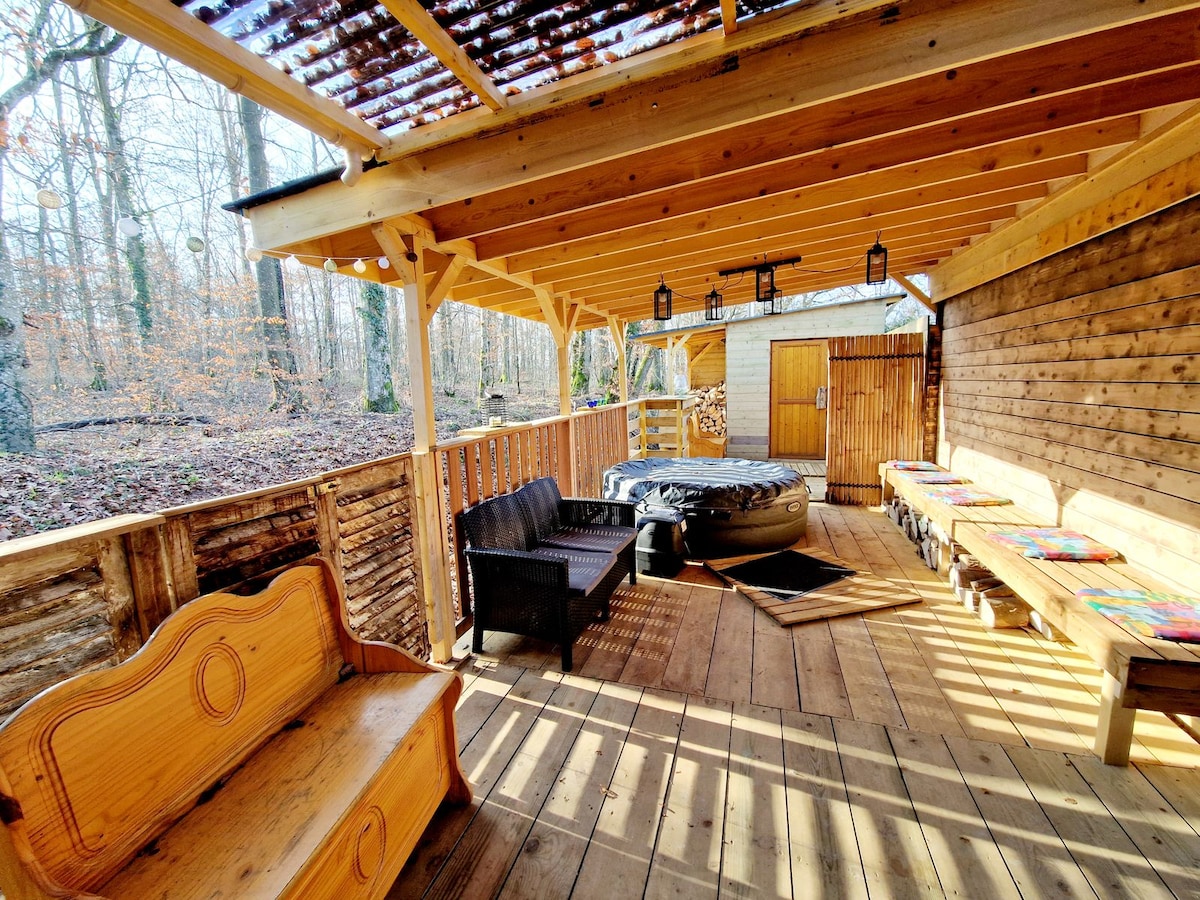 An outdoor seating area features wooden benches and a lounge chair under a rustic roof. A portable hot tub is positioned nearby, with log storage visible in the background. The surrounding forest offers a serene natural setting with trees and soft sunlight filtering through.