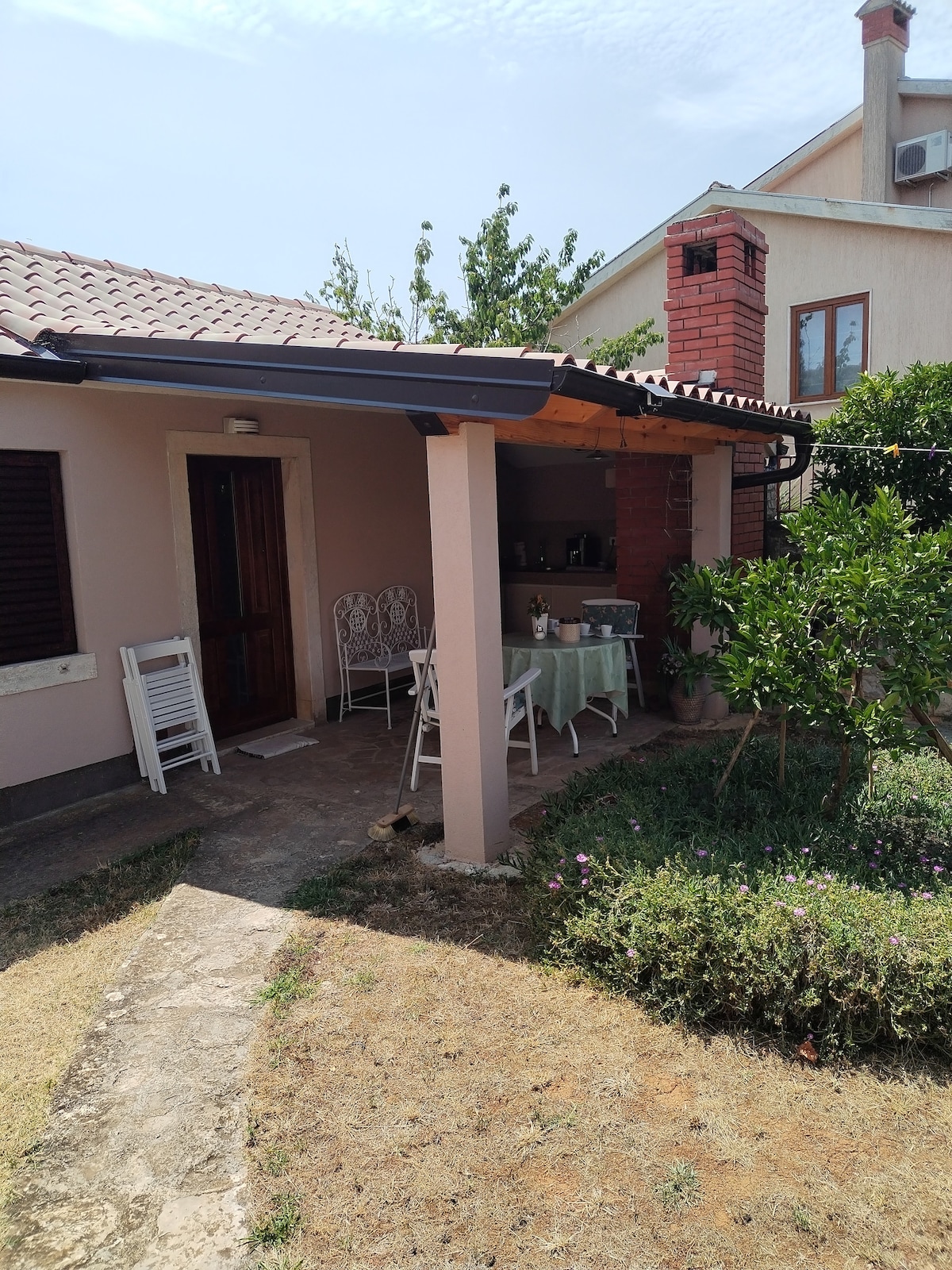 A front view of the cottage reveals a covered terrace with a dining table set for outdoor meals. The entrance features a wooden door, framed by white chairs and greenery. Stone pathways blend seamlessly with the surrounding landscaping, enhancing the inviting ambiance of the space.