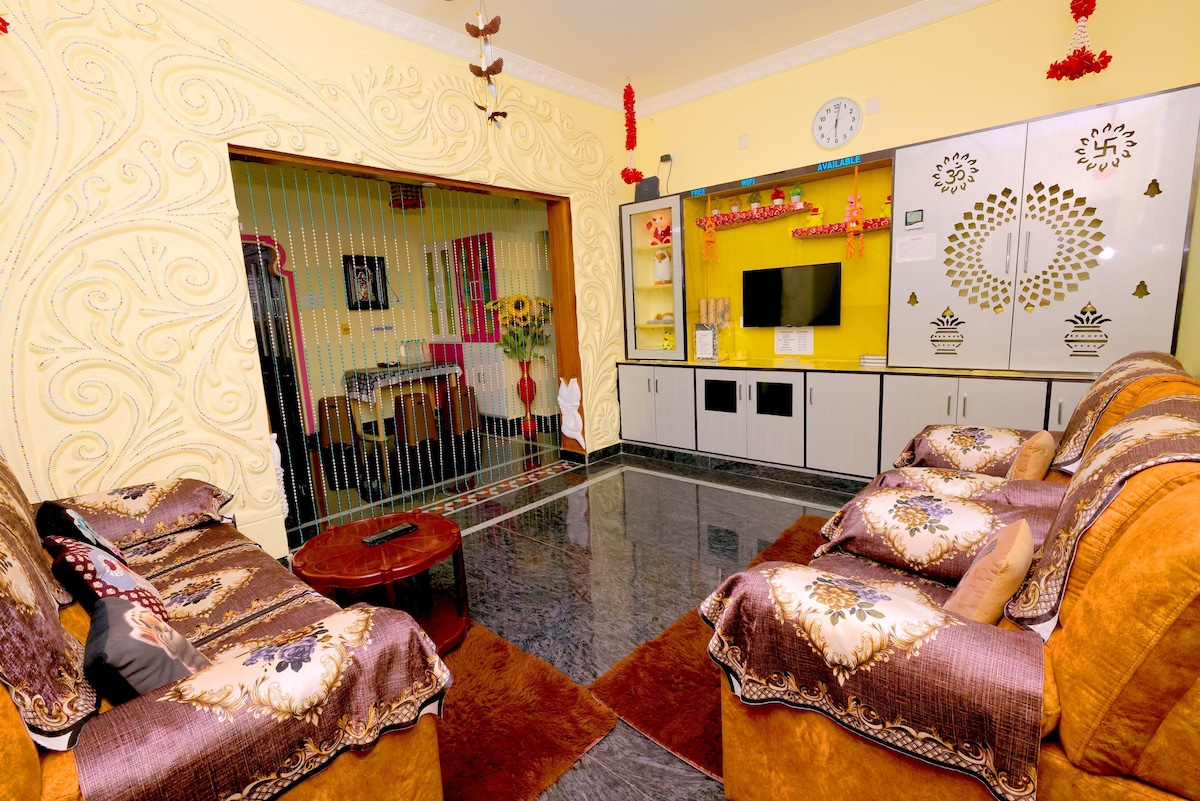 A living area features plush seating arrangements upholstered in earthy tones, accompanied by decorative cushions. A vibrant wall with intricate patterns frames a mounted television. The floor is highlighted by a deep color rug, complementing the bright yellow walls adorned with wall art.