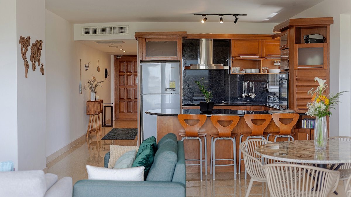 An open-plan living area is showcased, featuring a well-equipped kitchen with wood cabinetry and bar seating. Natural light floods the space, illuminating a cozy seating arrangement and a round dining table adorned with flowers. The decor reflects a modern and comfortable ambiance.