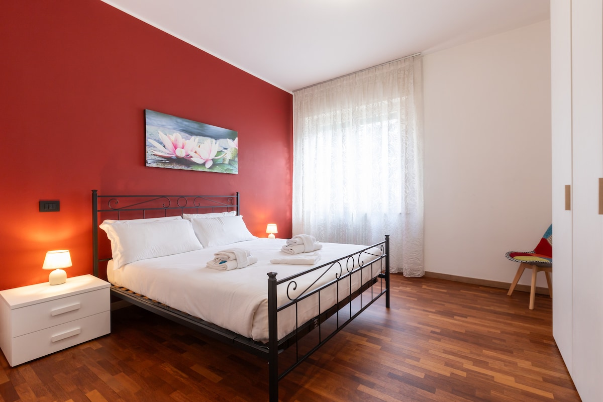 A bright bedroom features a double bed with white linens and two neatly arranged towels atop. A colorful artwork adorns a red wall, while natural light filters through a sheer curtain covering a large window. A nightstand with a lamp is positioned beside the bed.