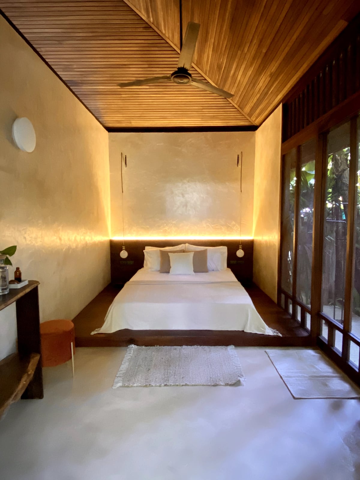 A spacious and serene sleeping area features a minimalist design with warm, wood accents. Soft lighting highlights the comfortable bed, while large windows allow for natural light and a view of the surrounding greenery.