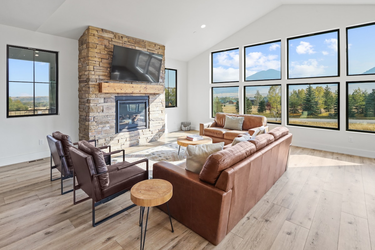 A bright living room is highlighted by large windows offering mountain views. A stone fireplace serves as a focal point, flanked by a mounted TV. Comfortable leather sofas and armchairs are arranged with a circular wooden table, enhancing the inviting space.