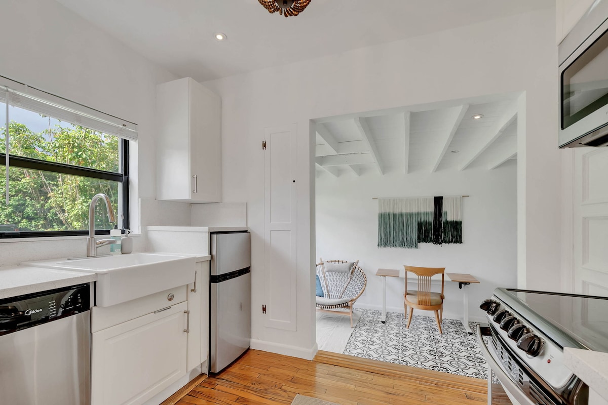 A modern kitchen is seen featuring white cabinetry, a stainless-steel sink, and an oven. Natural light flows in through a large window, illuminating the hardwood floor. Adjacent, a cozy dining area is visible with a light wood table and woven lounge chair set against a textured wall.