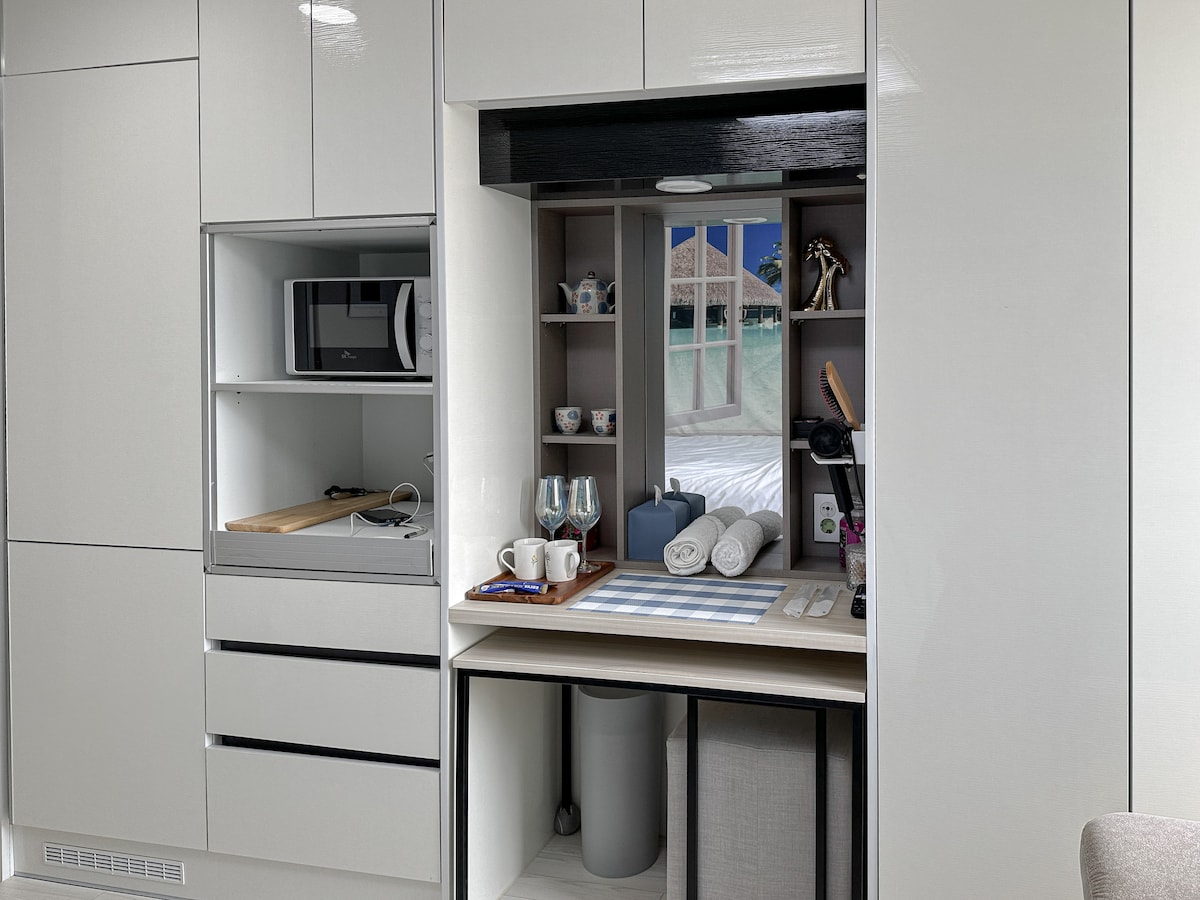 A modern kitchenette area features sleek cabinets and a built-in workspace. A microwave and a wooden cutting board are visible alongside neatly arranged cups and decorative items. A window view is framed, adding depth to the space. Clean towels are placed for convenience.