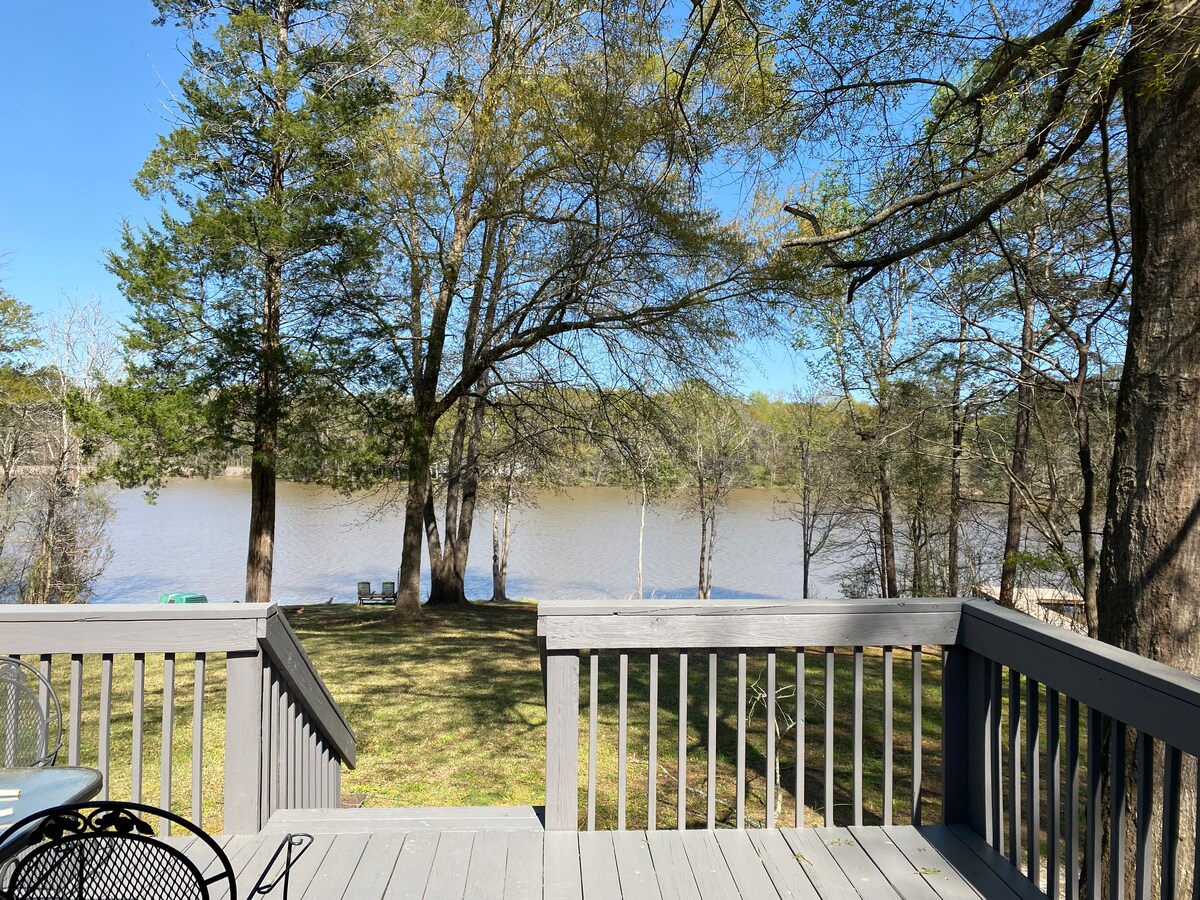 A peaceful view of a private lake is captured from the deck, framed by tall trees. A grassy area leads to the water's edge, with outdoor seating visible in the distance. Clear blue skies provide a bright backdrop to the tranquil scene.