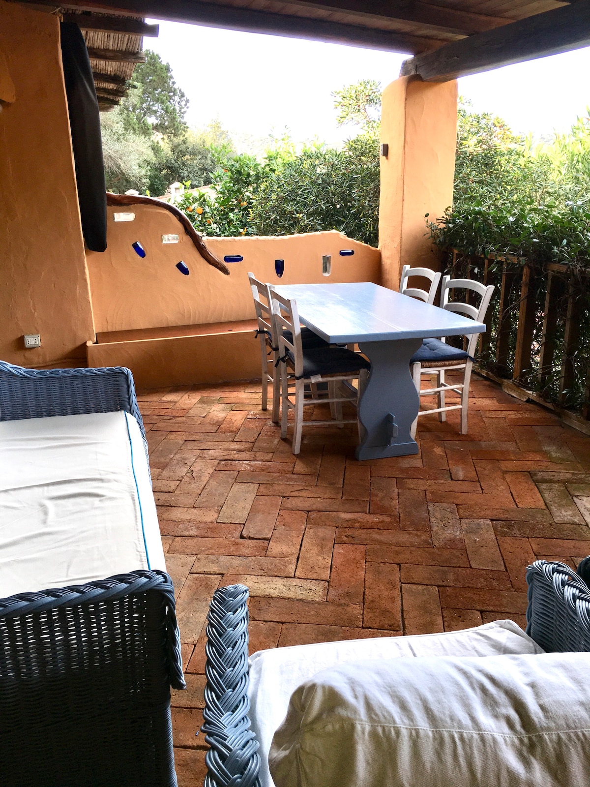 An outdoor veranda features a rectangular dining table surrounded by four chairs. Comfortable seating is provided on a nearby sofa. The space is enclosed by natural greenery, with a rustic wall accentuating the charming setting.