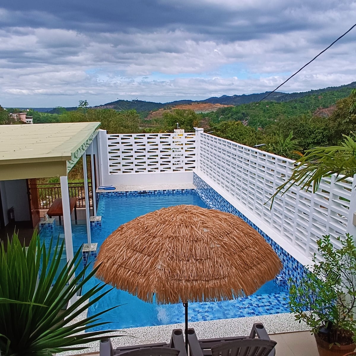 A glass swimming pool is surrounded by a serene landscape featuring distant mountains. An umbrella provides shade over the seating area, with lush greenery framing the scene. A covered space beside the pool offers a table for gatherings, emphasizing the inviting outdoor atmosphere.