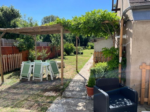 A shaded dining area features a wooden pergola draped with greenery. Four light-colored chairs are neatly arranged around a table. A pathway leads to a spacious garden, bordered by a low wooden fence and filled with grass and young plants.