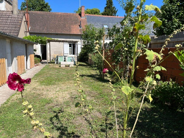 A spacious garden area is visible, featuring a grassy lawn bordered by flowering plants. In the background, the house can be seen, with a patio set available for outdoor dining and relaxation. The garden is enclosed, providing privacy and a sense of tranquility.
