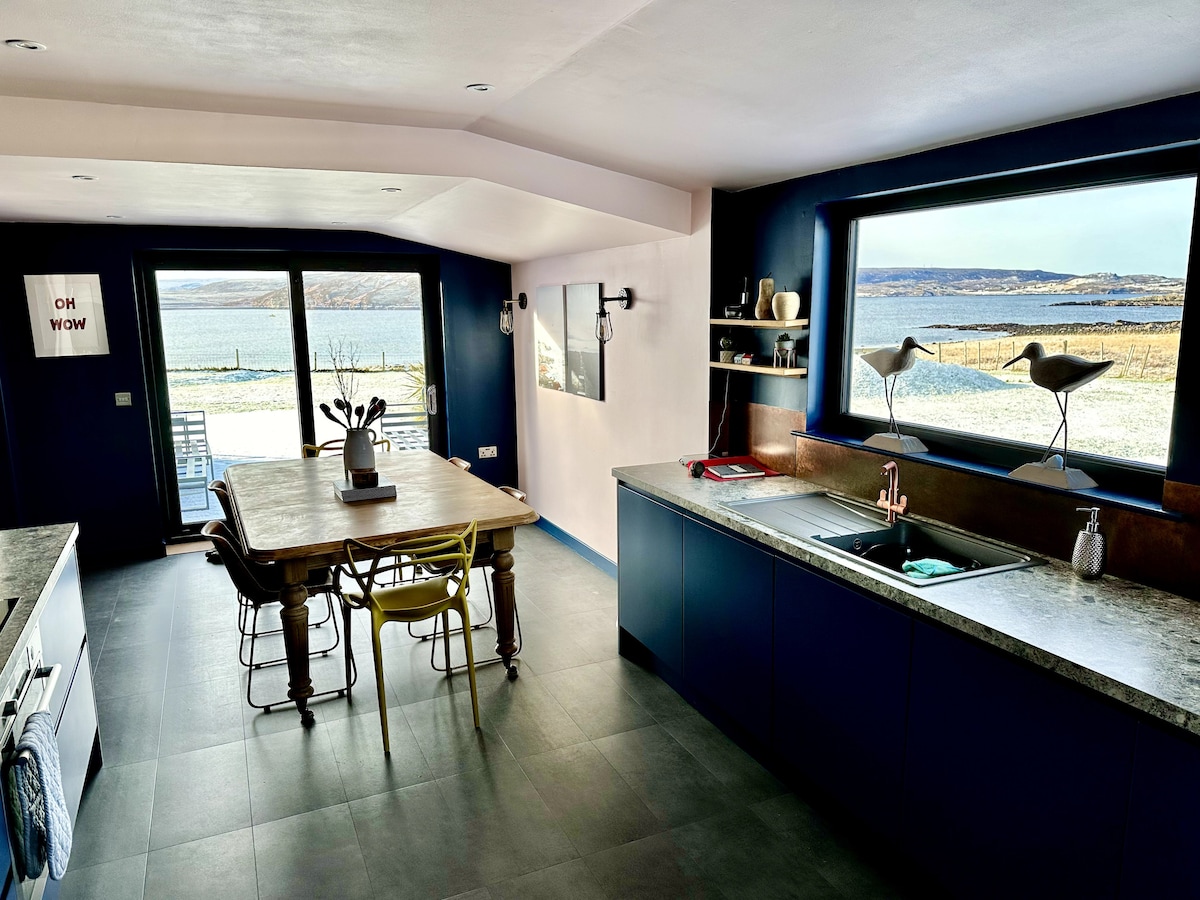 A spacious kitchen and dining area features modern appliances and an inviting dining table surrounded by a mix of colorful chairs. Large windows provide an unobstructed view of Loch Ewe, enhancing the connection between indoor and outdoor spaces.
