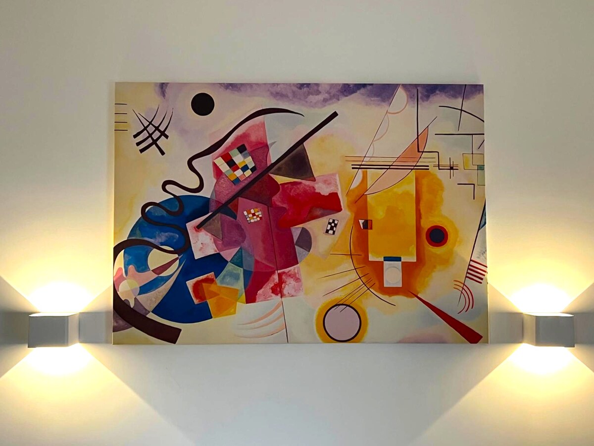An abstract painting dominates the wall, featuring a blend of warm and cool colors. Geometric shapes and flowing lines create a vibrant visual experience, flanked by two subtle wall sconces that provide gentle illumination.