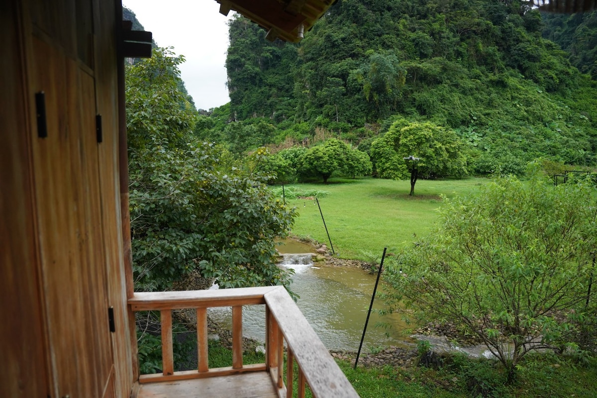 A wooden balcony offers a view of a lush, green garden featuring various trees. A stream flows gently through the landscape, while a distant hillside is covered in dense vegetation, creating a serene and natural setting.