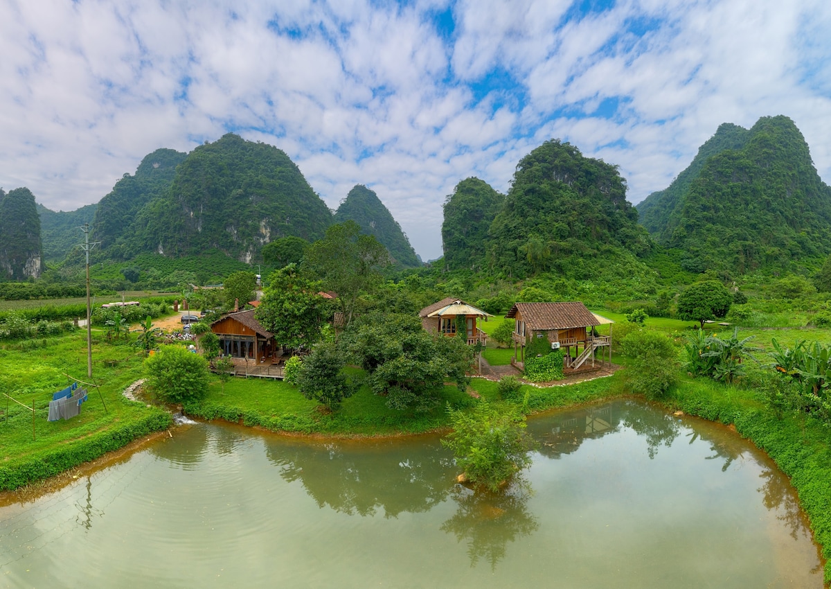 The image showcases Phong Sơn Retreat set amidst lush greenery and rolling mountains. Accompanied by a serene pond, the retreat features traditional-style buildings blending with nature, surrounded by rich vegetation under a partly cloudy sky.