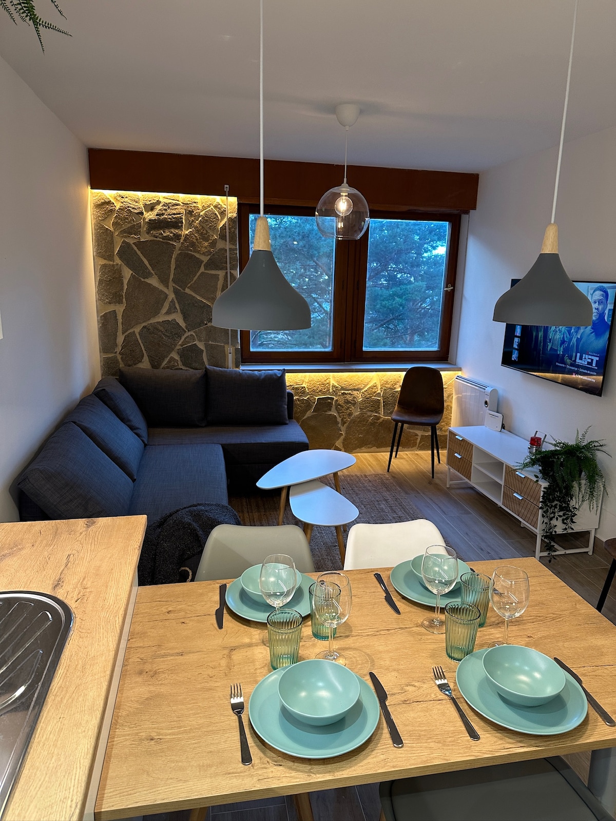 An open-concept living area features a modern sofa facing a flat-screen TV mounted on the wall. A light wood dining table is set with tableware in calming shades of green. Natural light filters through the large window, enhancing the warm ambiance.