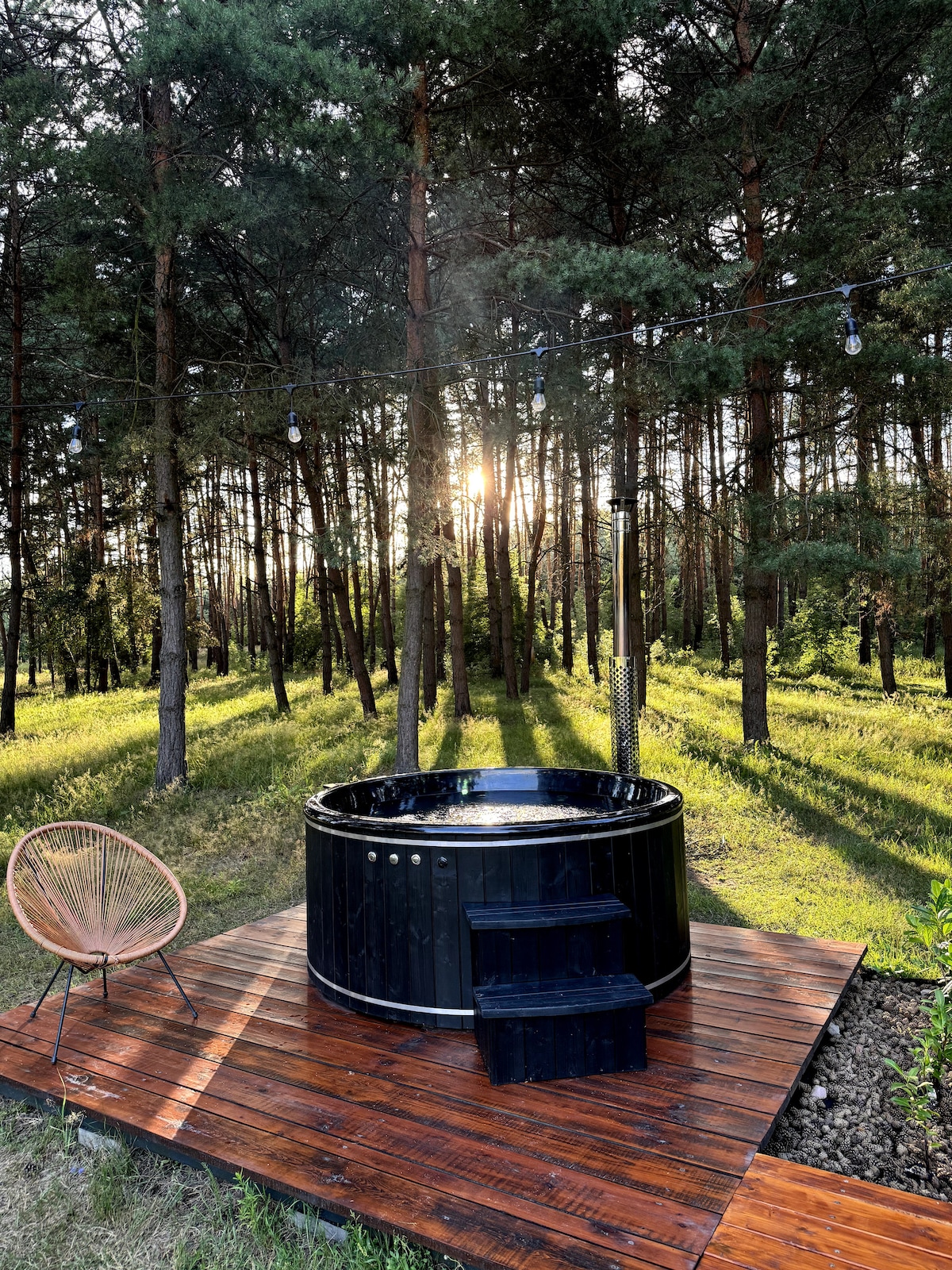 A wooden deck is featured with a black hot tub, surrounded by lush greenery and tall pine trees. Sunlight filters through the forest, casting soft shadows on the ground. An acapulco chair is positioned nearby, inviting relaxation.