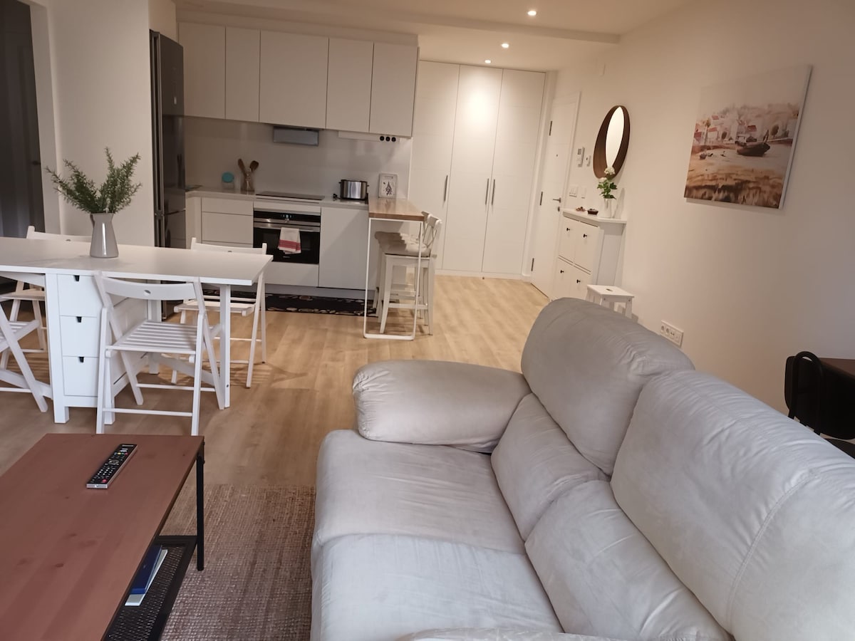 A modern kitchen and dining area are visible, featuring a white dining table with six chairs and a spacious gray sofa. Light wood flooring enhances the open layout, while ample natural light streams in from multiple sources. A neutral color palette is maintained throughout the space.