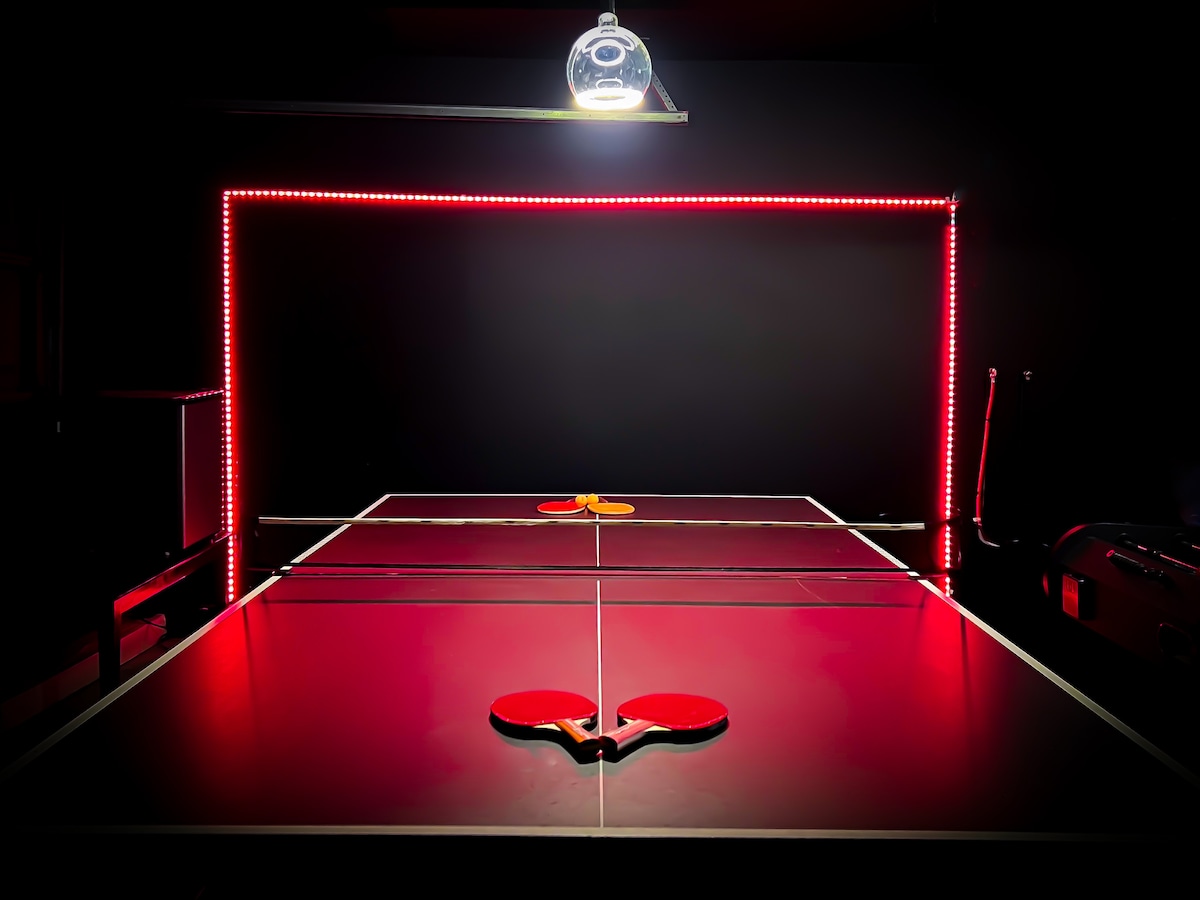 A well-lit ping pong table is highlighted by colorful LED lighting along the edge. Two paddles are positioned on the table, indicating readiness for play. The backdrop consists of a dark wall, creating a focused gaming environment.