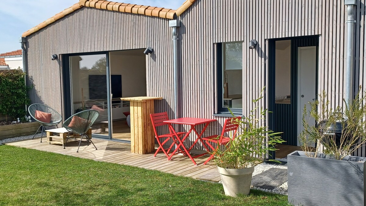 An outdoor terrace with wooden decking is shown, featuring a red table and chairs, complemented by lounge chairs. The smooth façade of the building is made of vertical wooden slats, and large glass doors open to the interior, surrounded by a green lawn and shrubs.