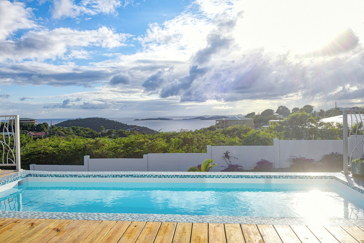 A sparkling private pool is framed by a wooden deck, offering a scenic view of lush greenery and expansive water. The horizon displays a blend of blue skies and clouds illuminated by sunlight, creating a serene atmosphere. The surrounding landscape showcases rolling hills and distant cays.