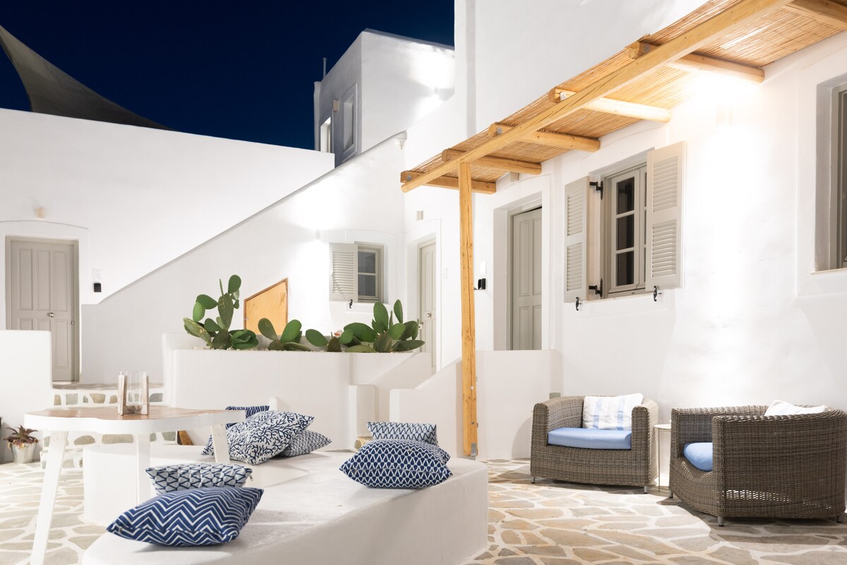 An outdoor patio area is framed by whitewashed walls and natural stone flooring. Comfortable seating arrangements feature wicker chairs and blue cushions. Potted plants add greenery, while overhead lighting creates an inviting atmosphere during the evening. The space maintains a minimalist and cozy vibe.